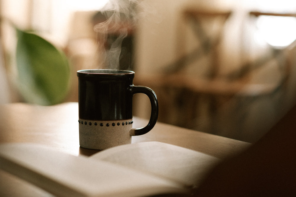  tasse noire et beige de café fumant avec un livre et verdure en arrière plan